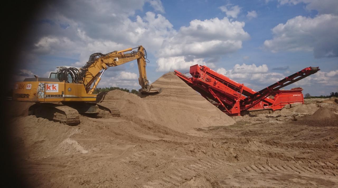 Żółta koparka Liebherr nabiera urobek z hałdy, obok czerwona przesiewarka na tle błękitnego nieba z chmurami.