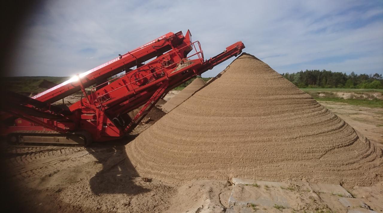 Czerwona przesiewaczka mobilna marki Terex Finlay przesypuje piasek na stożkowy pryzmę, widoczne ślady opon na piasku i drzewa w tle.
