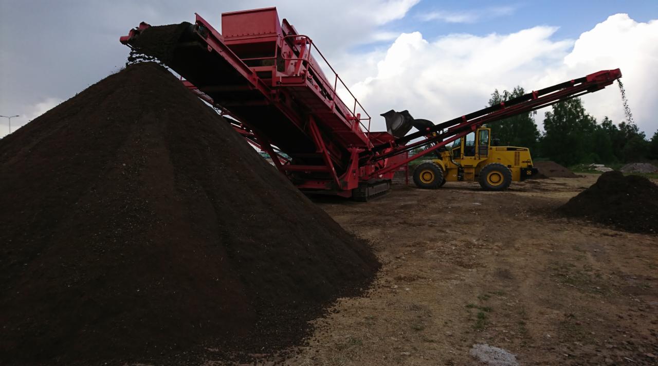 Czerwony przesiewacz taśmowy usypuje ciemną ziemię ogrodową na pryzmę, w tle żółta ładowarka kołowa i zieleń drzew.