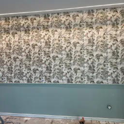 Ściana z tapetą w motywy tropikalne, oddzielona listwą od pomalowanej na jednolity, pastelowy kolor dolnej części, widoczny odkurzacz i resztki po pracy.