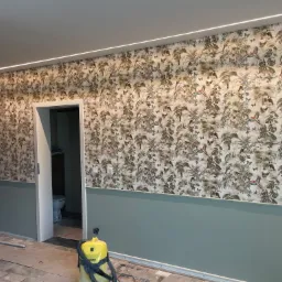 Pokój po remoncie z tapetą w tropikalne wzory, pomalowaną dolną częścią ściany na zielono i widocznym odkurzaczem w rogu, z otwartymi drzwiami do łazienki.