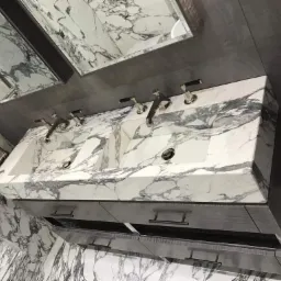 Wykończona łazienka z podwójną umywalką z białego marmuru z szarymi żyłami, zamontowaną na szarej szafce z metalowymi uchwytami. Na ścianach i podłodze marmur, nad umywalką lustro w ciemnej ramie.