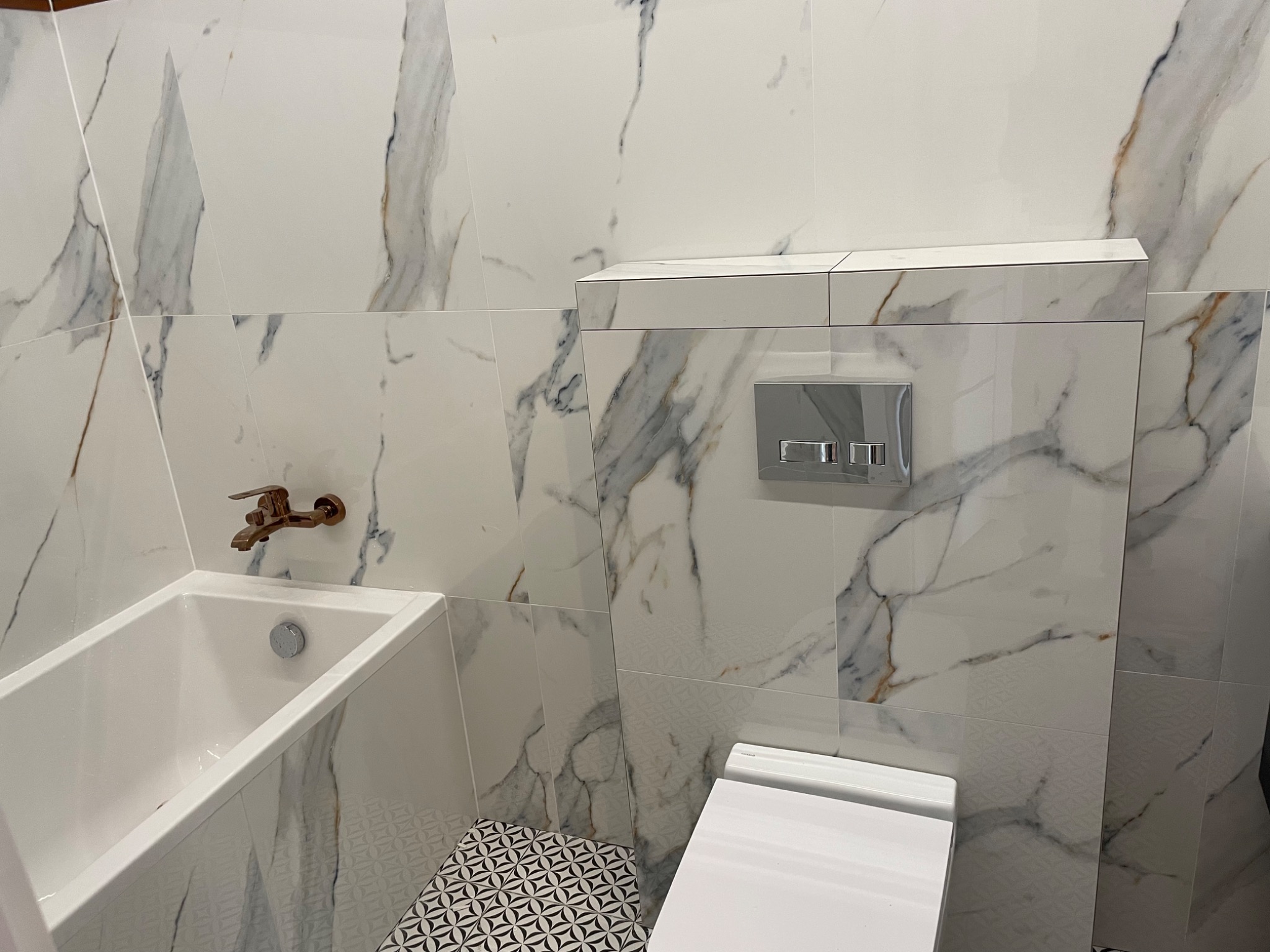 Wykończona łazienka z wanną, podwieszaną toaletą i płytkami imitującymi marmur z akcentami w kolorze złota, widoczna bateria wannowa w odcieniu różowego złota.