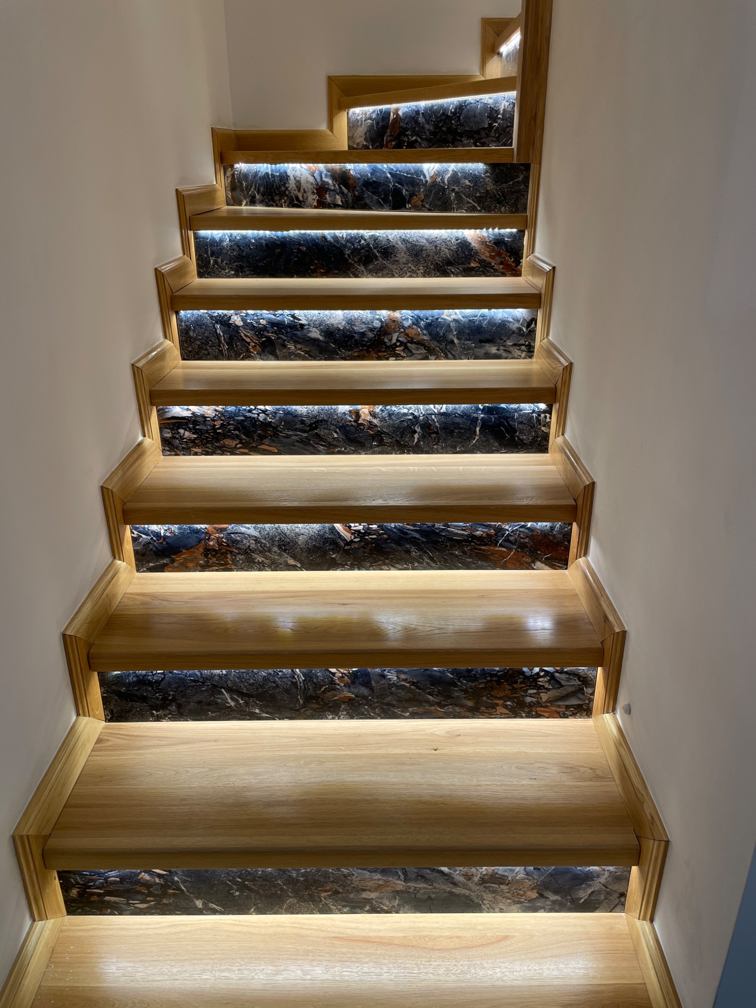 Schody drewniane z marmurowymi podstopnicami i subtelnym oświetleniem LED pod każdym stopniem, widok z dołu ukazujący eleganckie wykończenie.