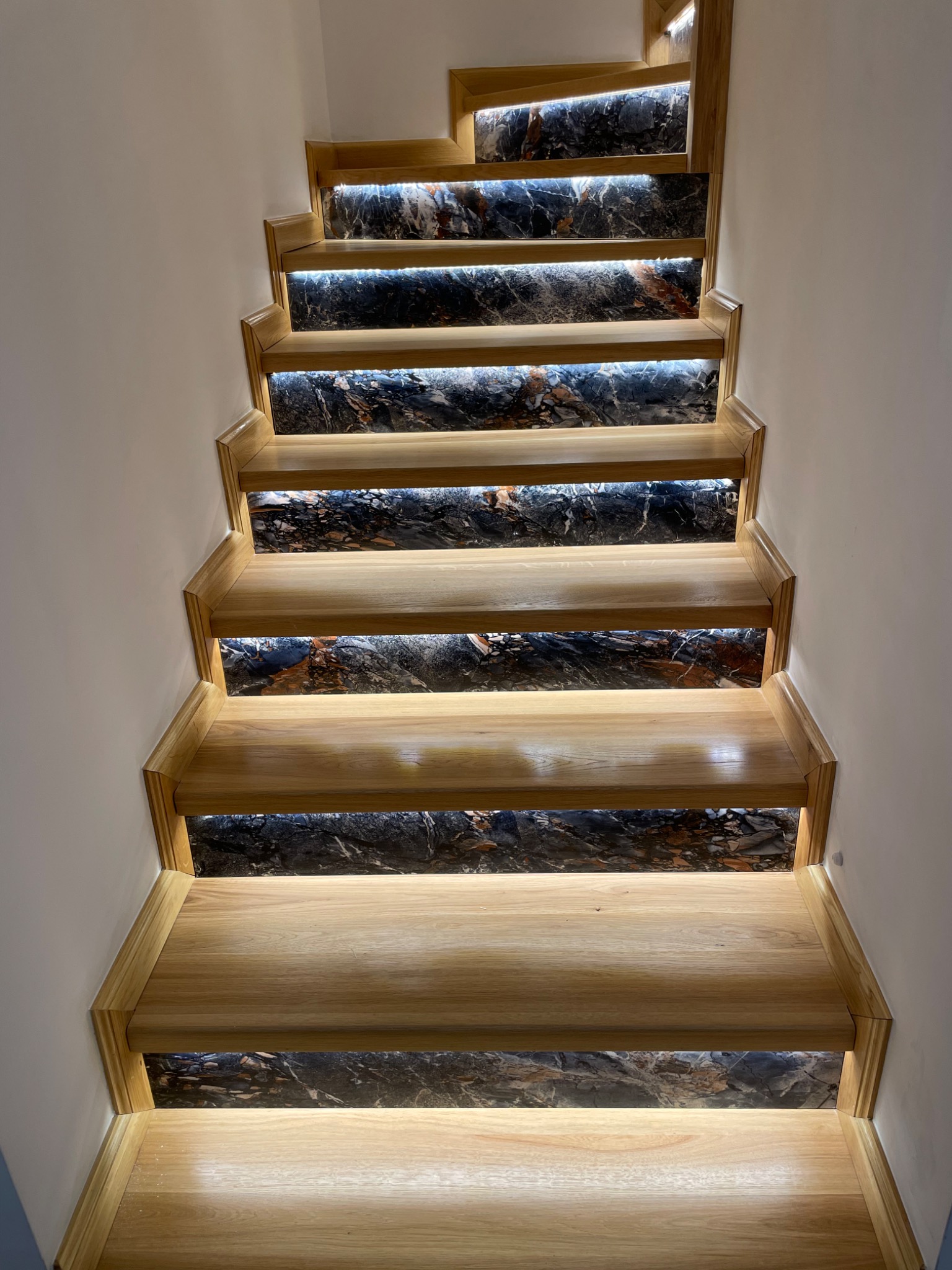 Drewniane schody z marmurowymi podstopnicami, podświetlone listwami LED, widok z dołu.