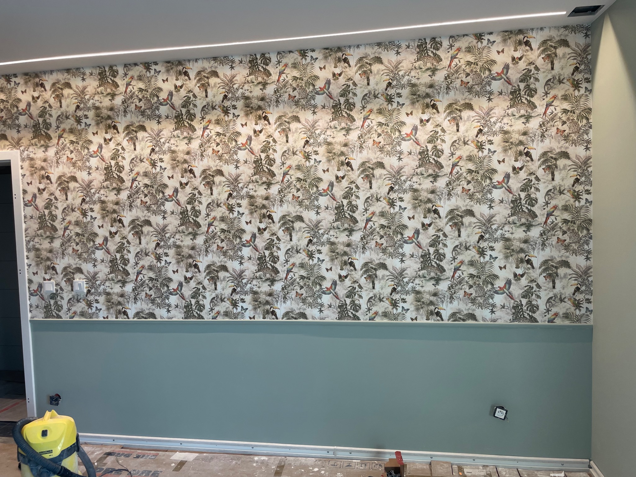 Ściana z tapetą w motywy tropikalne, oddzielona listwą od pomalowanej na jednolity, pastelowy kolor dolnej części, widoczny odkurzacz i resztki po pracy.