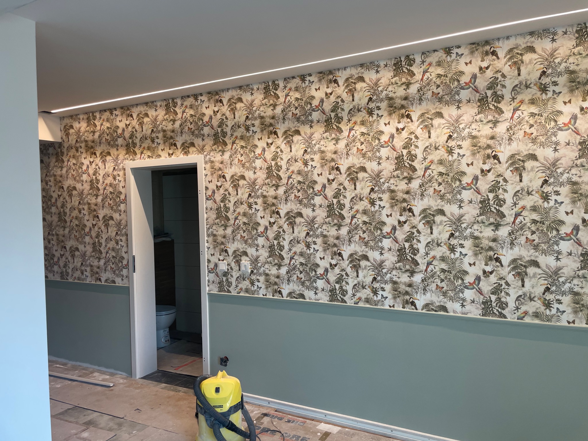 Pokój po remoncie z tapetą w tropikalne wzory, pomalowaną dolną częścią ściany na zielono i widocznym odkurzaczem w rogu, z otwartymi drzwiami do łazienki.