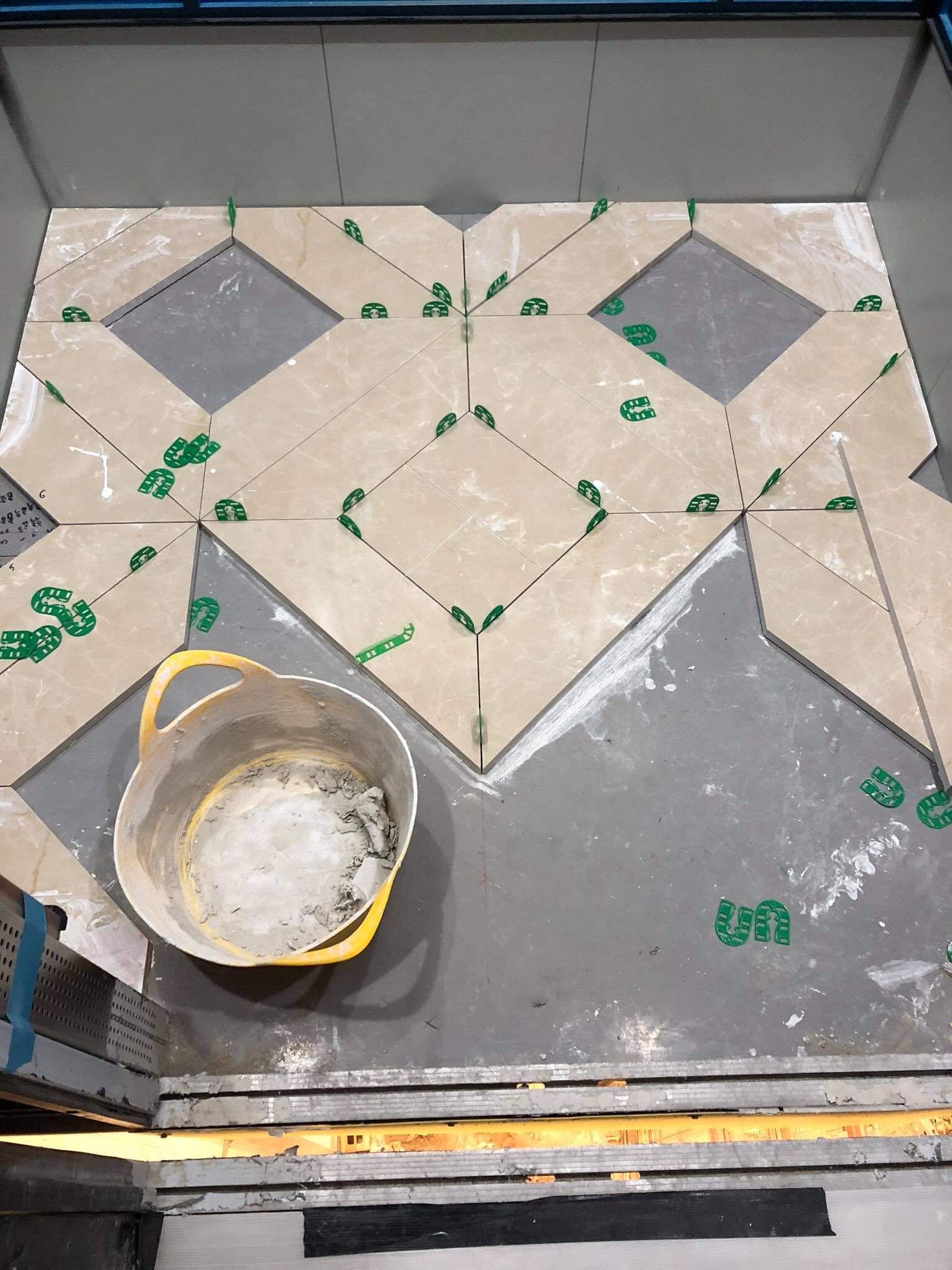 Układanie beżowych płytek podłogowych w geometryczny wzór z użyciem zielonych klinów dystansowych, widoczny żółty kubeł z zaprawą.