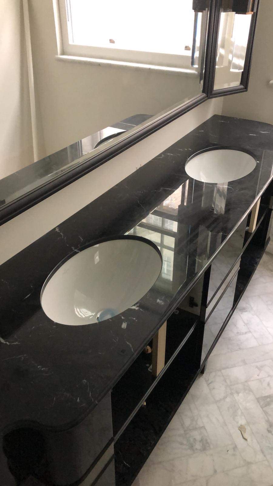 Czarny marmurowy blat z dwiema owalnymi umywalkami w łazience, widoczne odbicie okna w blacie, białe marmurowe płytki podłogowe.