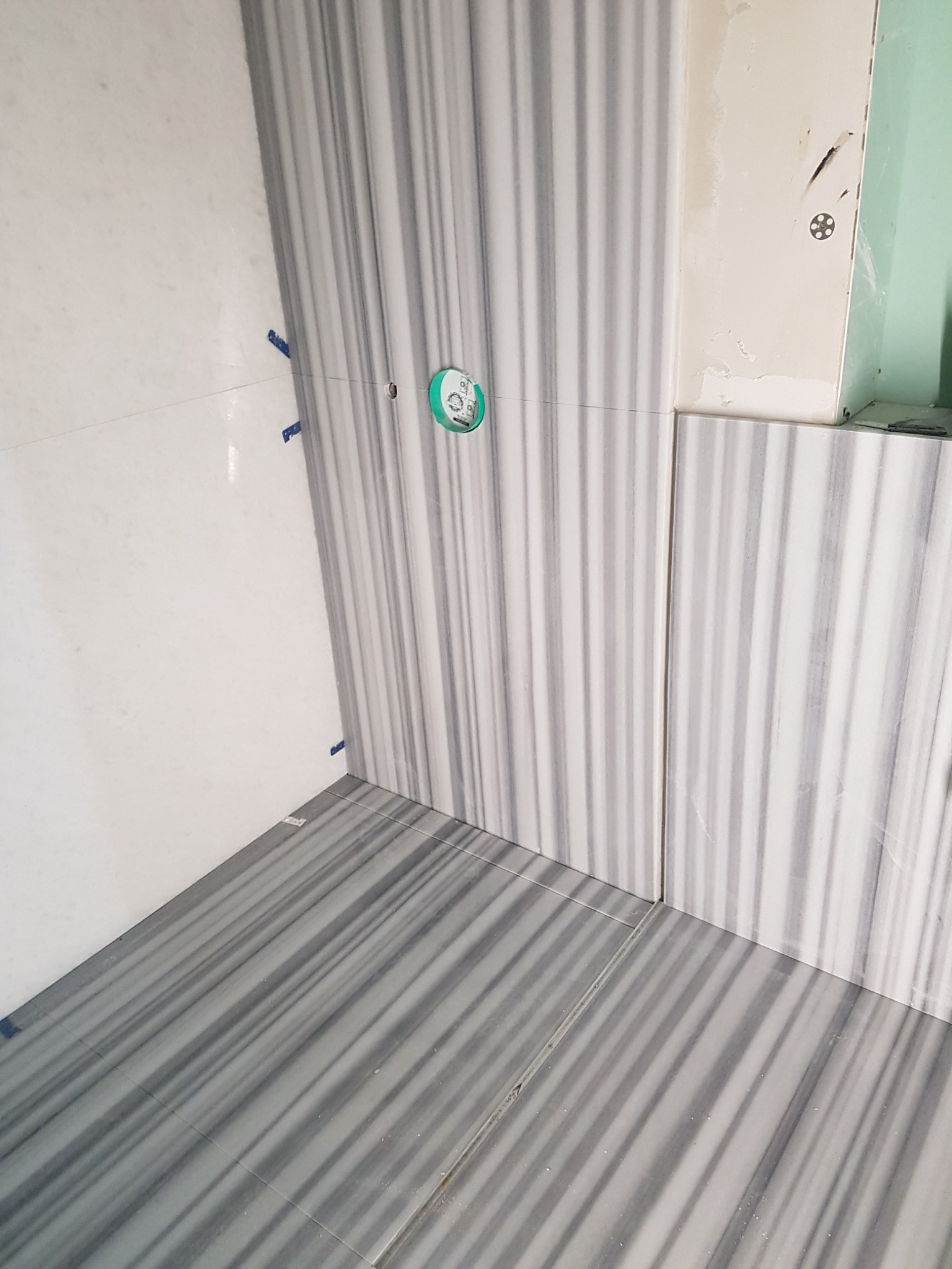 Ułożone płytki podłogowe i ścienne z szarym wzorem w paski; niebieskie kliny dystansowe widoczne w spoinach; otwór pod instalację wodną.