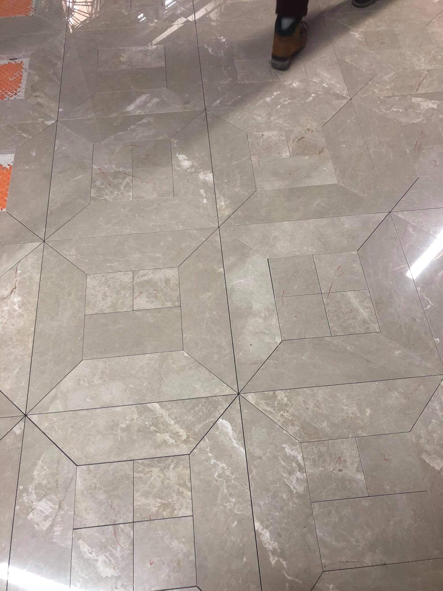 Ułożona podłoga z płytek w odcieniach beżu i szarości, z wzorem geometrycznym, widoczne fragmenty podkładu i buta pracownika w rogu.