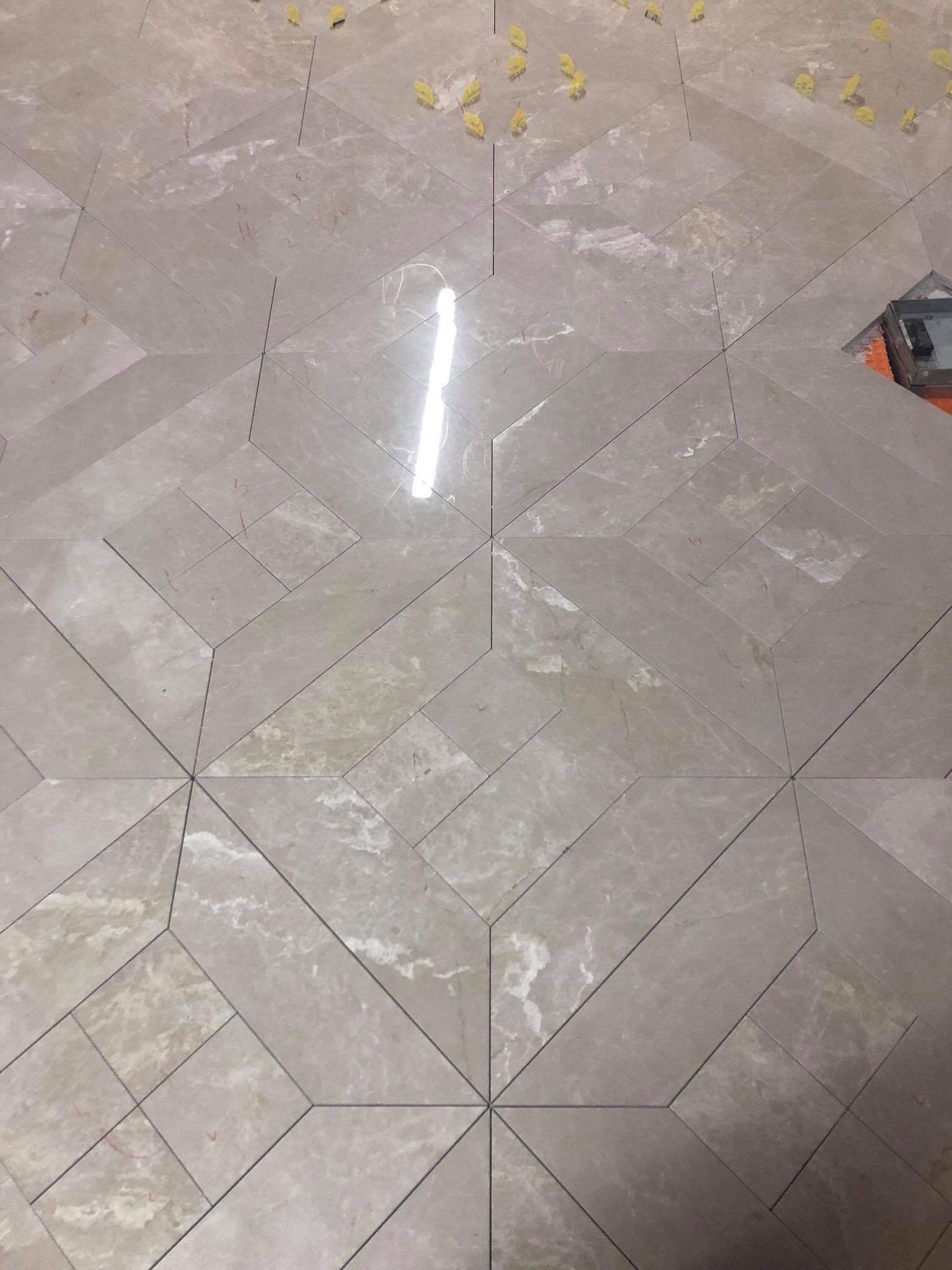 Ułożona pod kątem podłoga z szarych płytek w geometryczny wzór, z widocznymi żółtymi podkładkami dystansowymi i fragmentem narzędzia do cięcia płytek w prawym górnym rogu.