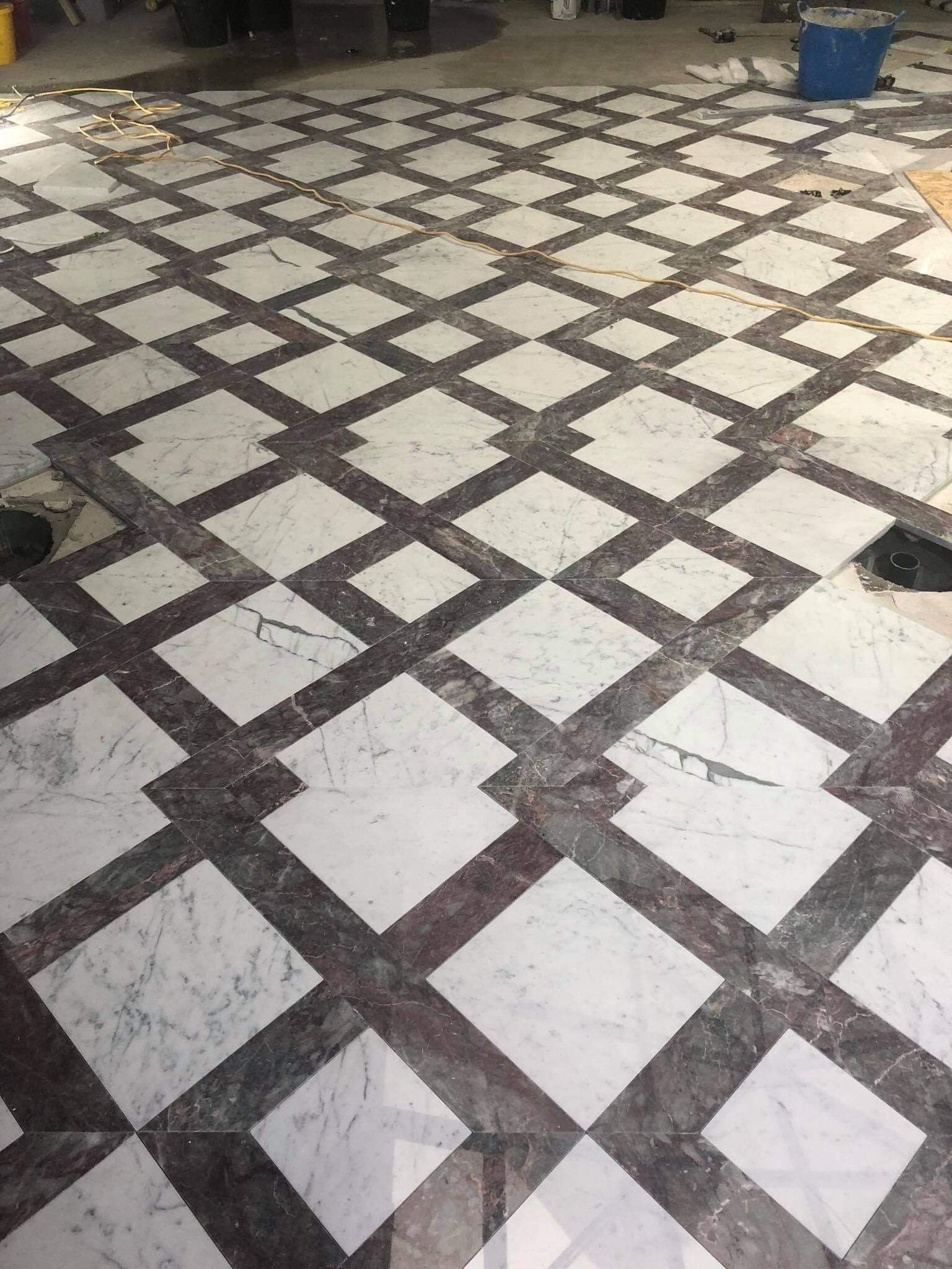 Wzorzysta podłoga z białych i ciemnych marmurowych płytek ułożonych w romby, widoczne niedokończone fragmenty posadzki i instalacja w Rokicinach.