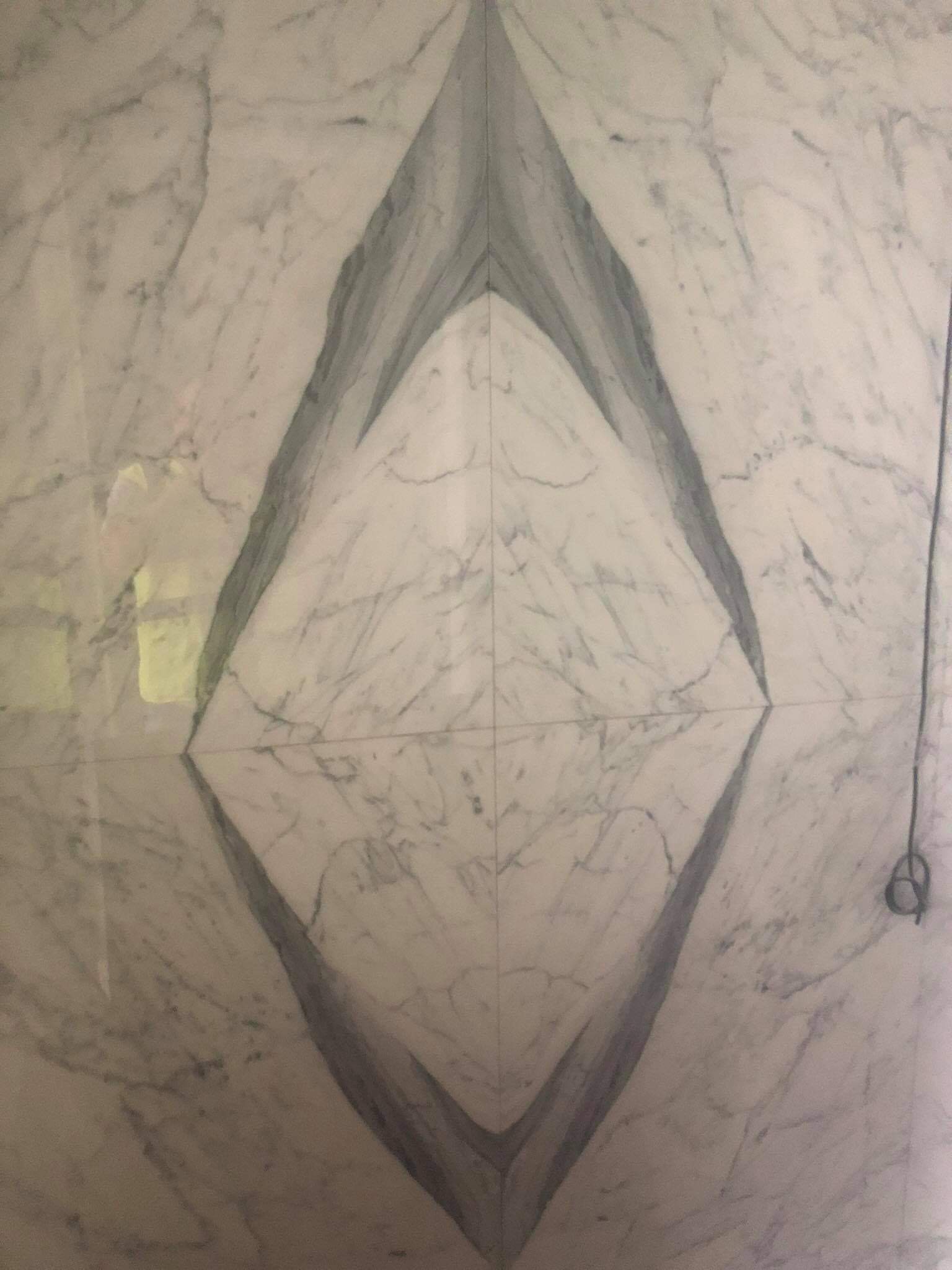 Położona na ścianie biała marmurowa płytka z szarymi żyłami tworzącymi symetryczny wzór w kształcie rombu, widoczne połączenia między płytkami.