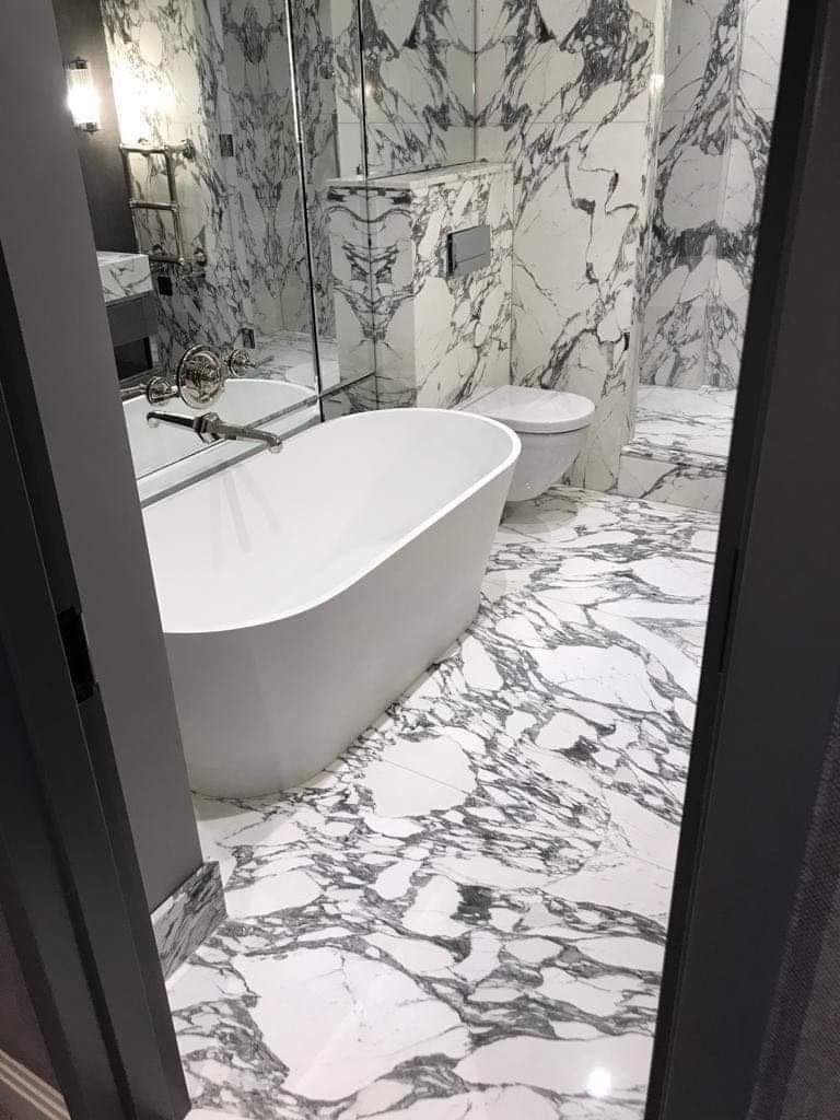 Nowoczesna łazienka wykończona białym marmurem z czarnymi żyłami, widok na wolnostojącą wannę, toaletę i umywalkę z lustrem, całość oświetlona subtelnym światłem.