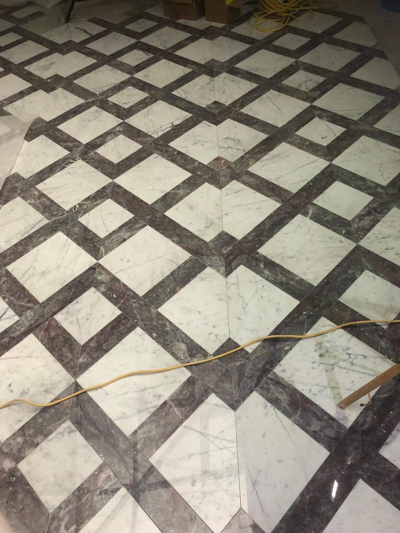 Marmurowa podłoga ułożona we wzór rombów z białych i szarych płytek, widoczny fragment żółtego kabla i nogi stołu.