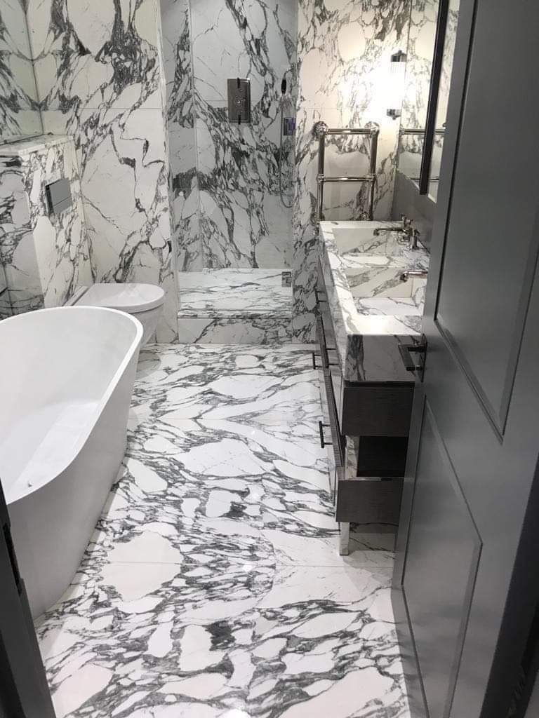 Elegancka łazienka wykończona białym marmurem z czarnymi żyłkami, z wolnostojącą wanną, kabiną prysznicową i szafką z umywalką; widoczne otwarte szuflady.