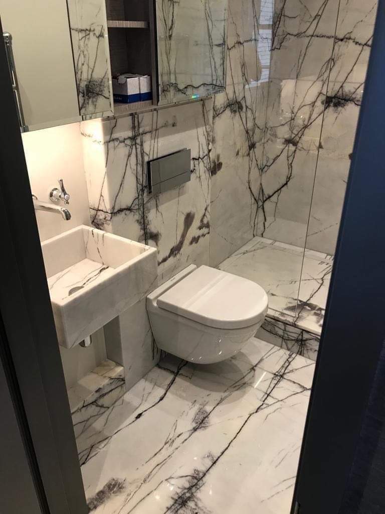 Nowoczesna łazienka z białymi płytkami imitującymi marmur z czarnymi żyłkami, wisząca toaleta i umywalka z tego samego materiału, szklana kabina prysznicowa w tle.