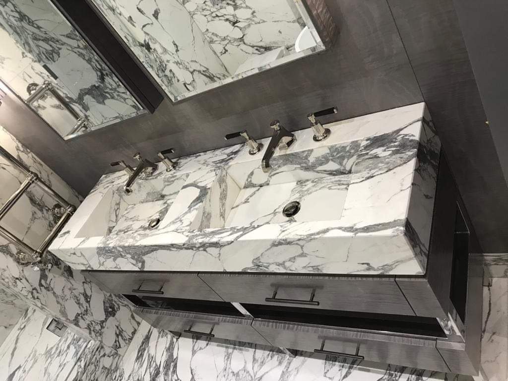 Wykończona łazienka z podwójną umywalką z białego marmuru z szarymi żyłami, zamontowaną na szarej szafce z metalowymi uchwytami. Na ścianach i podłodze marmur, nad umywalką lustro w ciemnej ramie.