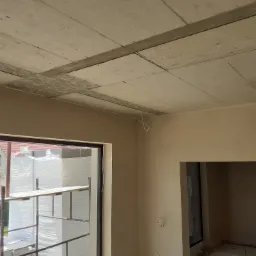 Surowy betonowy strop w nowym budynku z widocznymi śladami szalunków, ściany pokryte świeżym tynkiem w odcieniu beżu, widoczne wystające przewody elektryczne, przez okno widoczne rusztowanie...