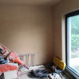 Świeżo położone tynki maszynowe w Lubinie, widoczna maszyna do tynkowania, drabina i narzędzia w pomieszczeniu z dużym oknem.