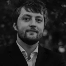 Portret adwokata w czarnym garniturze i białej koszuli, z wzorzystą poszetką, na rozmytym tle zieleni.