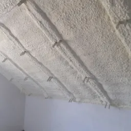 Poddasze ocieplone białą pianką natryskową z widocznymi metalowymi uchwytami mocującymi, surowy stan wykończenia ścian i podłogi.