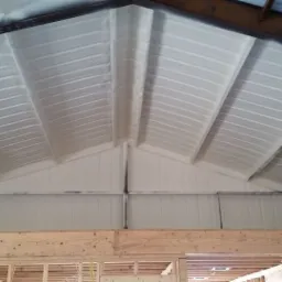 Poddasze z metalową konstrukcją dachu pokryte warstwą białej pianki izolacyjnej, widoczne drewniane elementy konstrukcyjne ściany.