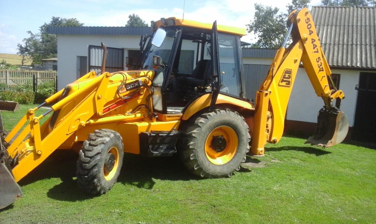 Żółta koparko-ładowarka JCB 3CX stoi na trawniku przed budynkiem gospodarczym z szarą blachodachówką, widoczny numer telefonu na ramieniu koparki.