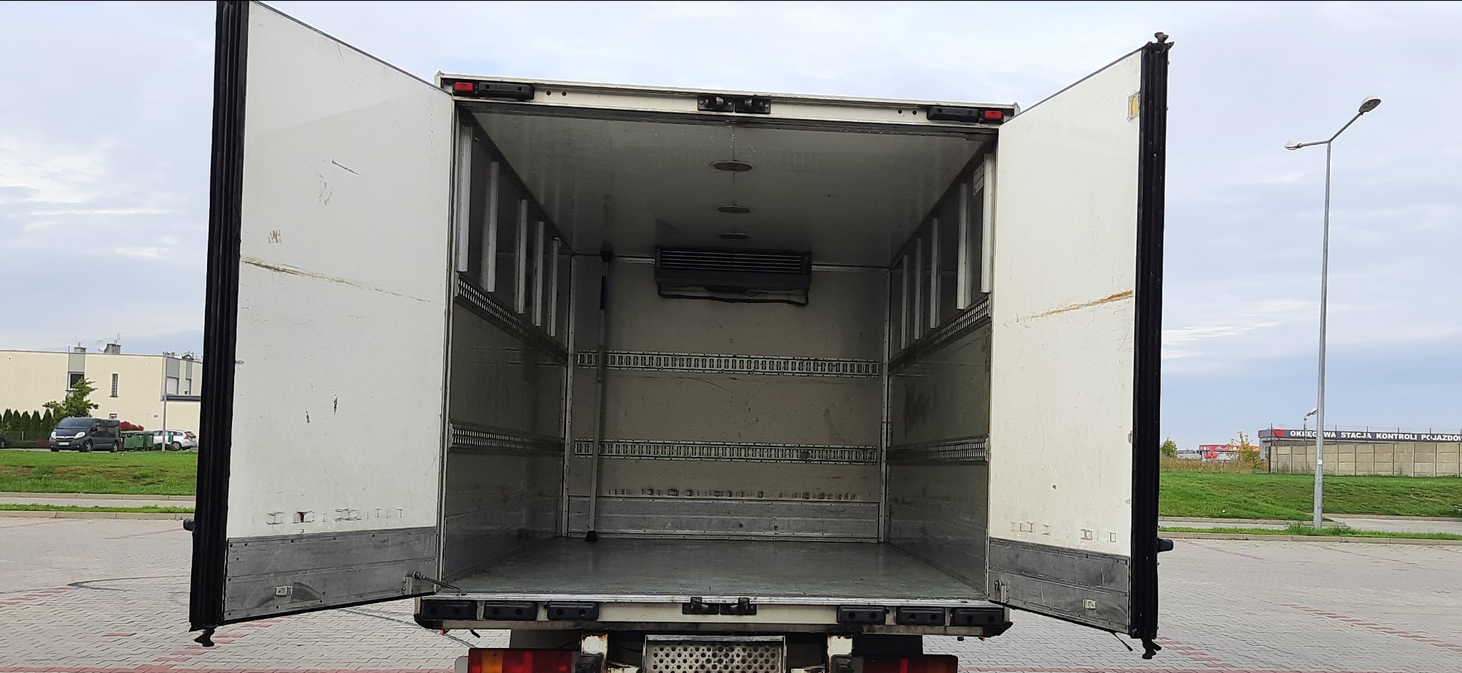 Wnętrze pustej chłodni samochodu ciężarowego z otwartymi drzwiami, widoczne szyny mocujące ładunek i agregat chłodniczy.