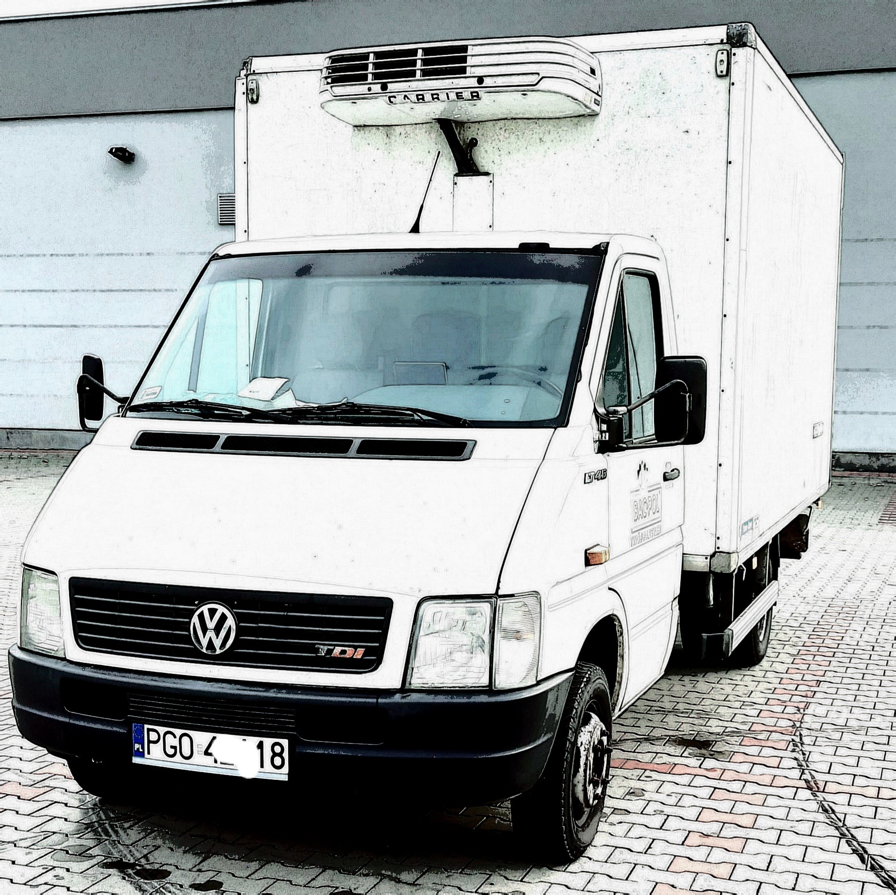 Biały samochód dostawczy z chłodnią Carrier na dachu, zaparkowany na kostce brukowej przed budynkiem.