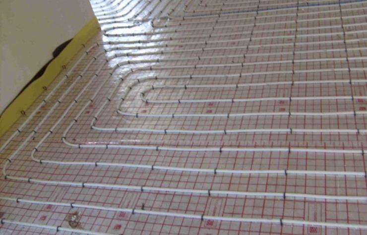 Białe rurki ogrzewania podłogowego ułożone równolegle na podłożu z siatką w kolorze czerwonym i brązowym, przymocowane czarnymi uchwytami.