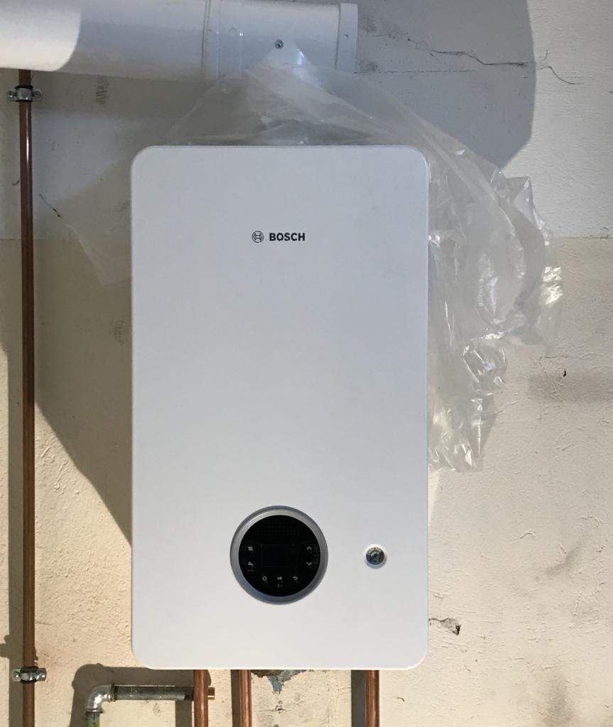 Biała pompa ciepła Bosch zamontowana na szarej ścianie, z widocznymi rurami miedzianymi i folią ochronną na górze, instalacja w trakcie lub tuż po montażu.