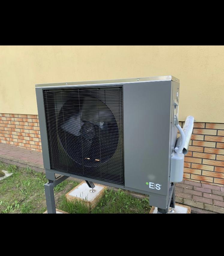 Szara pompa ciepła zamontowana na zewnątrz budynku z żółtą elewacją i fragmentem ceglanej ściany, widoczny wąż karbowany i logo firmy na obudowie.