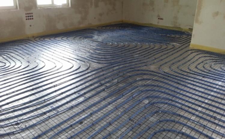 Niebieskie rury ogrzewania podłogowego ułożone w spiralny wzór na podkładzie, widoczne w surowym stanie ściany i okna w tle.