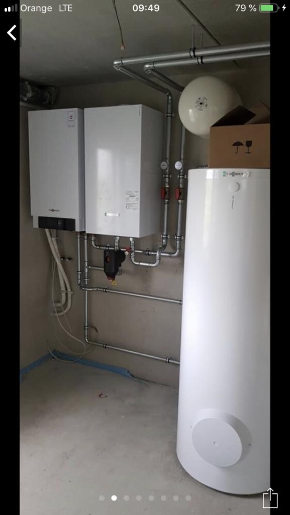 Dwa białe kotły grzewcze Viessmann zamontowane na szarej ścianie, z widocznymi rurami, filtrem i zasobnikiem ciepłej wody. Częściowo widoczny karton z symbolami delikatnego obchodzenia się...