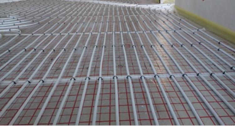 Białe rury ogrzewania podłogowego ułożone na siatce z czerwonymi kwadratami, przymocowane czarnymi uchwytami, widok z góry.