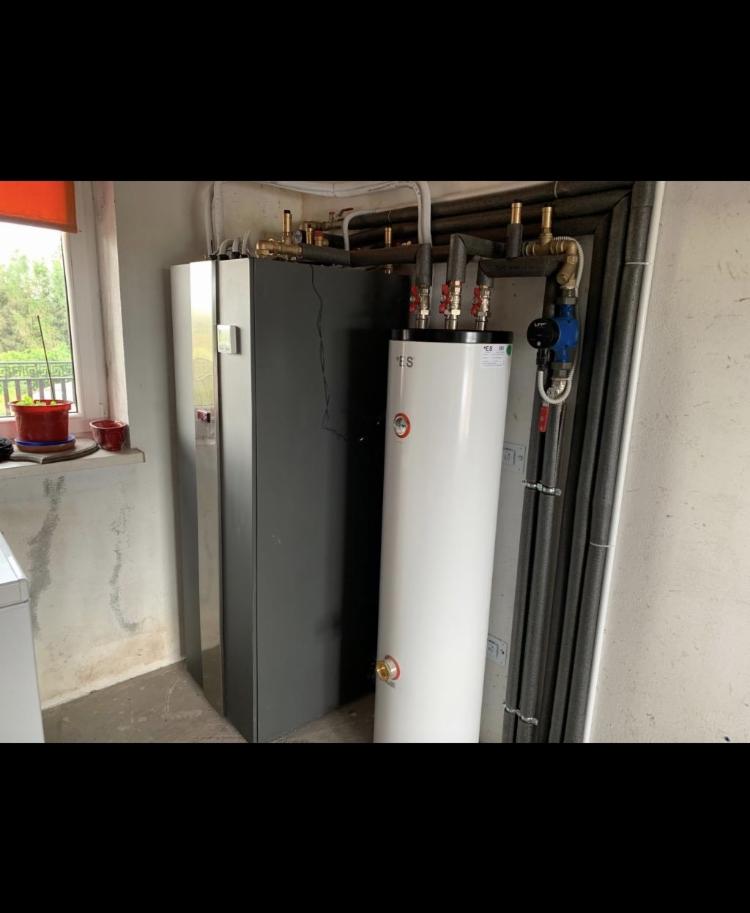 Kompaktowa instalacja grzewcza z szarą obudową i białym zbiornikiem, widoczne rury i armatura, zamontowana w pomieszczeniu z oknem.