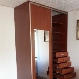 Brązowa szafa wnękowa z lustrem i otwartymi półkami z szufladami, widoczna na tle ściany z tapetą.