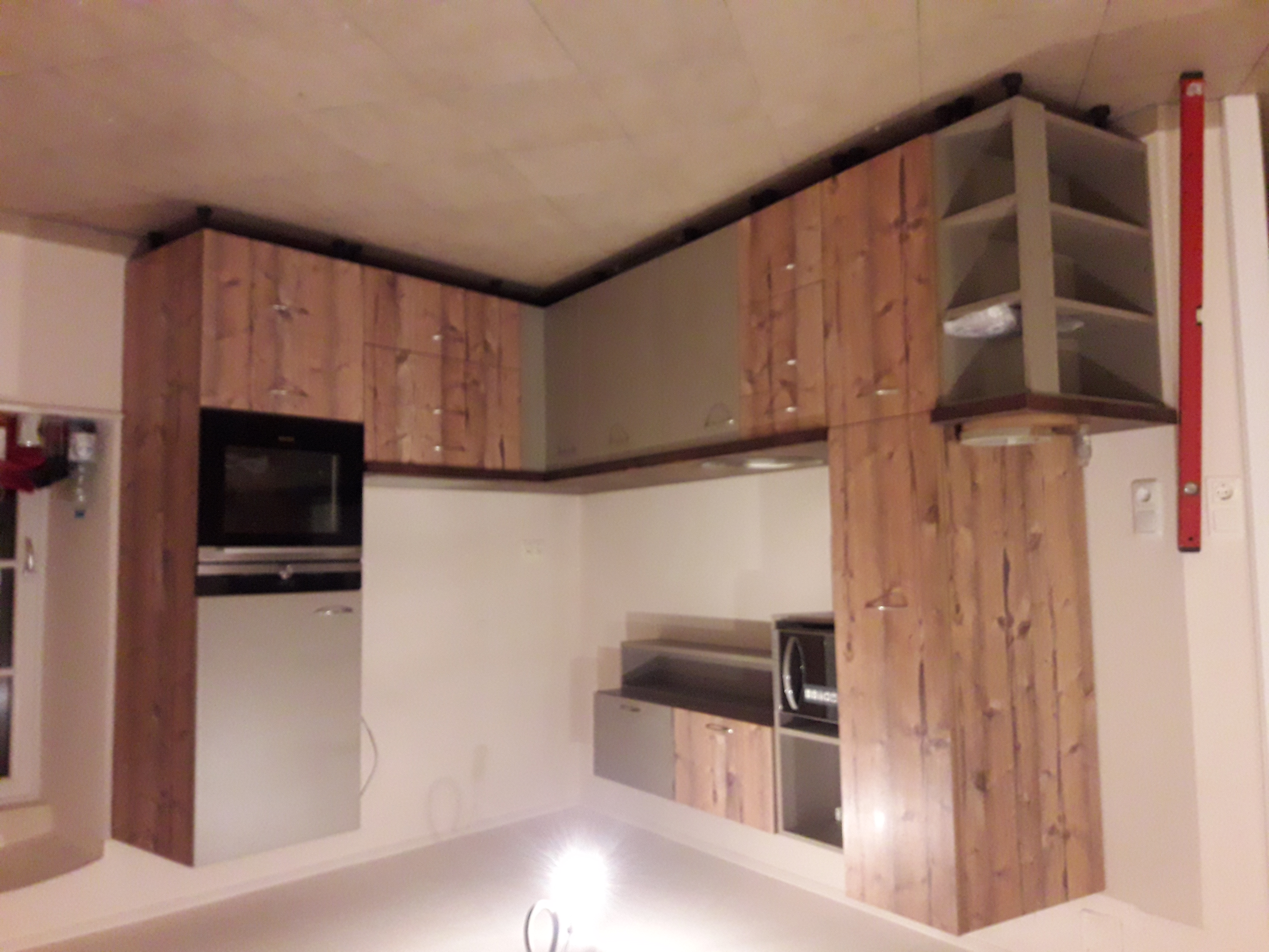 Nowoczesna kuchnia z drewnianymi frontami szafek, wbudowanym piekarnikiem i mikrofalówką, oświetlona lampą podłogową, z poziomnicą opartą o ścianę po prawej stronie.