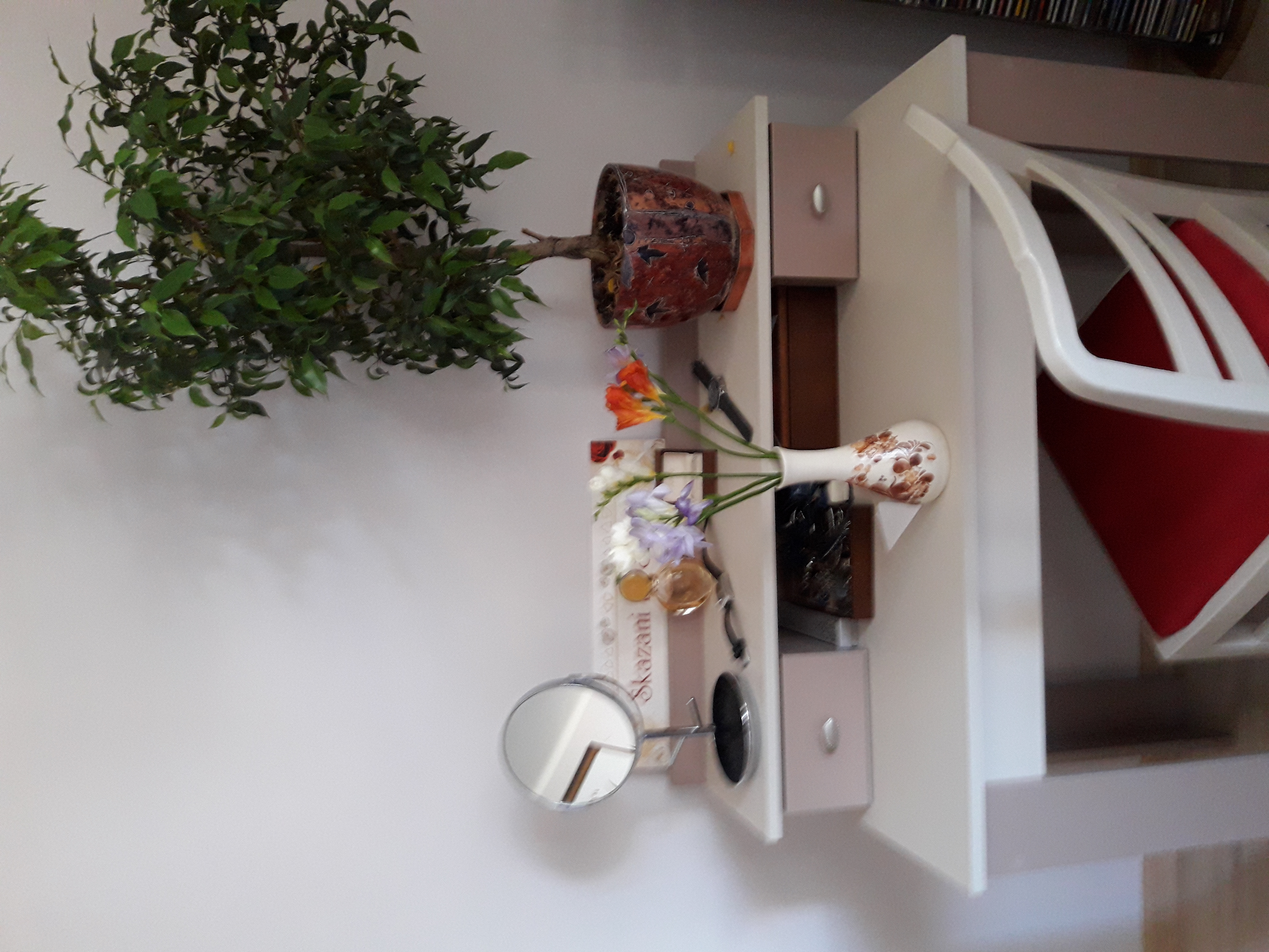 Fragment białej szafki nocnej z otwartą szufladą, na której stoi doniczka z kwiatami, wazon z bukietem i lusterko kosmetyczne, w tle czerwone krzesło.