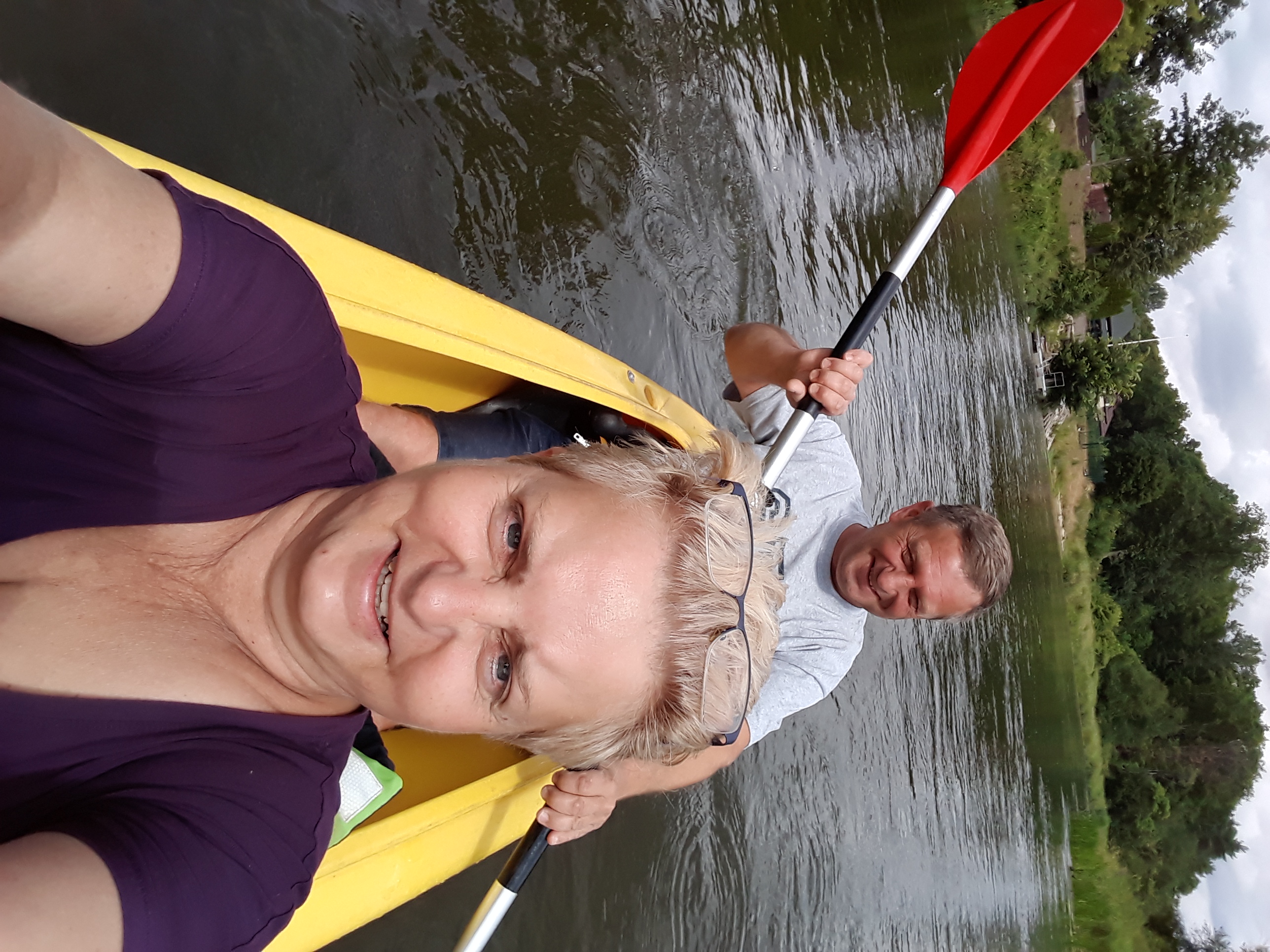 Selfie z kajaka: uśmiechnięta blondynka w fioletowej koszulce i mężczyzna wiosłujący czerwoną łopatką na tle zielonych brzegów rzeki.
