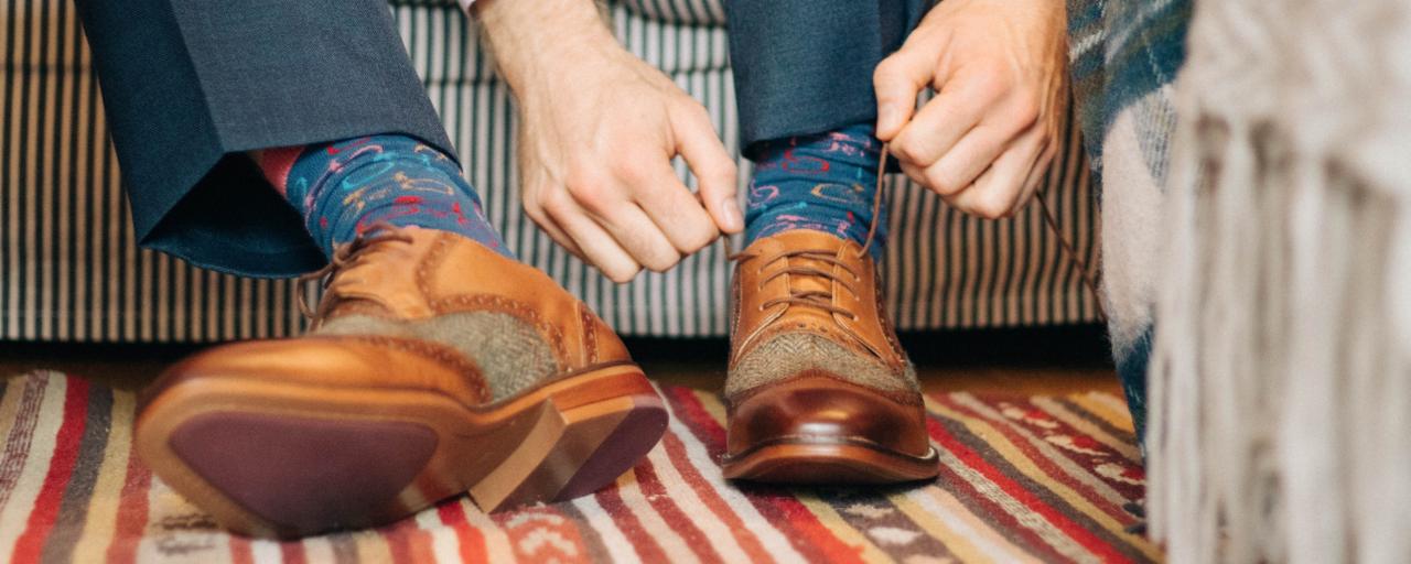 Mężczyzna w Gdańsku zawiązuje brązowe, skórzane buty z tweedowymi wstawkami, nosząc kolorowe skarpetki w geometryczne wzory, na wzorzystym dywanie.