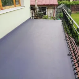 Szary, wykończony balkon z ozdobną, kutą balustradą na tle zielonego ogrodu i jasnej elewacji budynku.