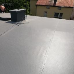 Jakub borecki - Szary, płaski dach pokryty papą termozgrzewalną z widocznym kominem wentylacyjnym i pracownikiem w oddali, na tle budynku z pomarańczową dachówką.