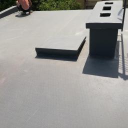 Jakub borecki - Szary, płaski dach pokryty papą termozgrzewalną z widocznymi kominami i pracownikiem w tle, wykonującym prace dekarskie.