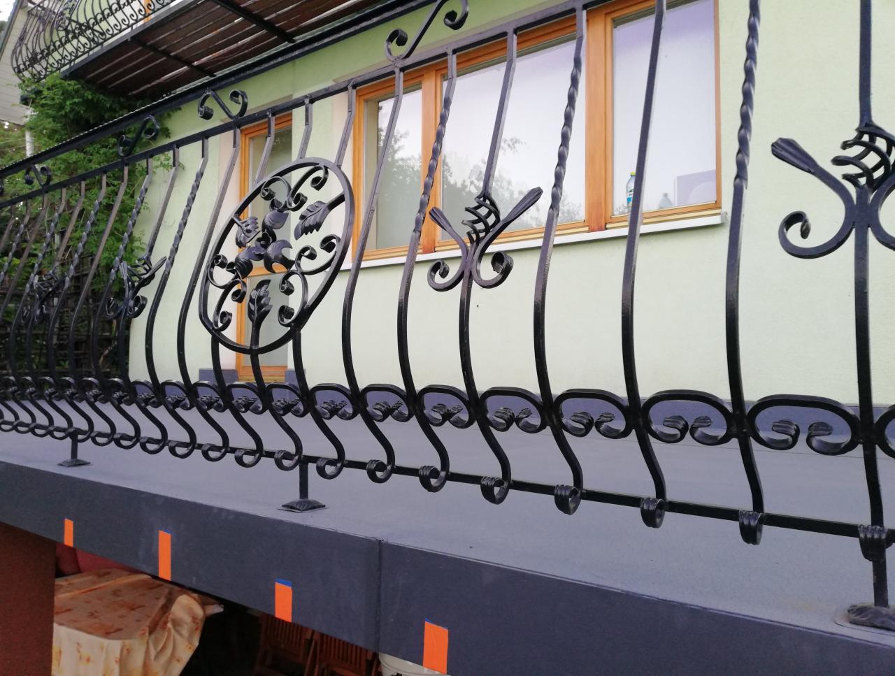 Kute, czarne balustrady balkonowe z motywami roślinnymi na tle jasnej elewacji i drewnianych okien.