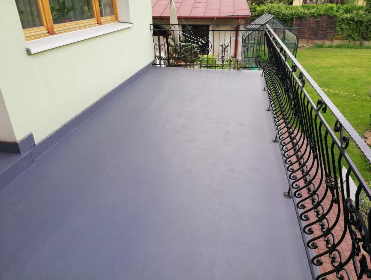 Szary, wykończony balkon z ozdobną, kutą balustradą na tle zielonego ogrodu i jasnej elewacji budynku.