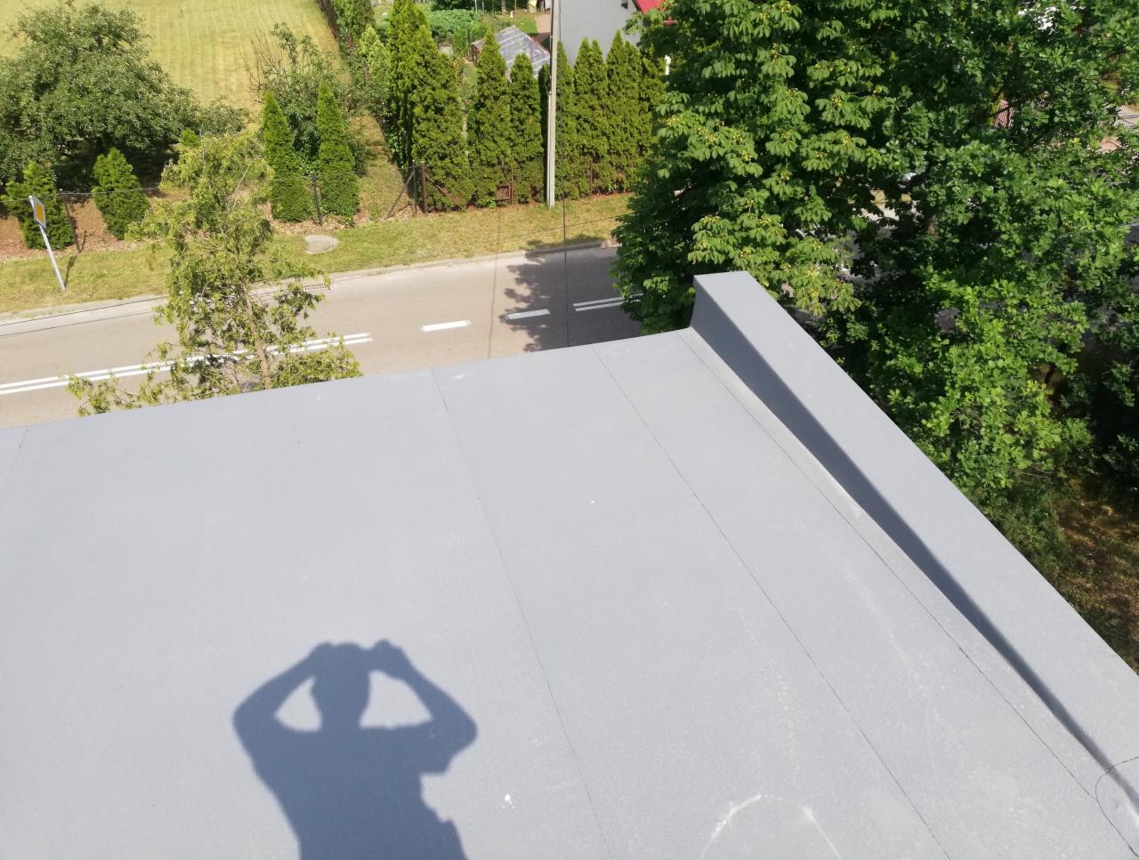 Szary, płaski dach pokryty papą termozgrzewalną, widoczna cień osoby robiącej zdjęcie, w tle ulica i zieleń drzew.