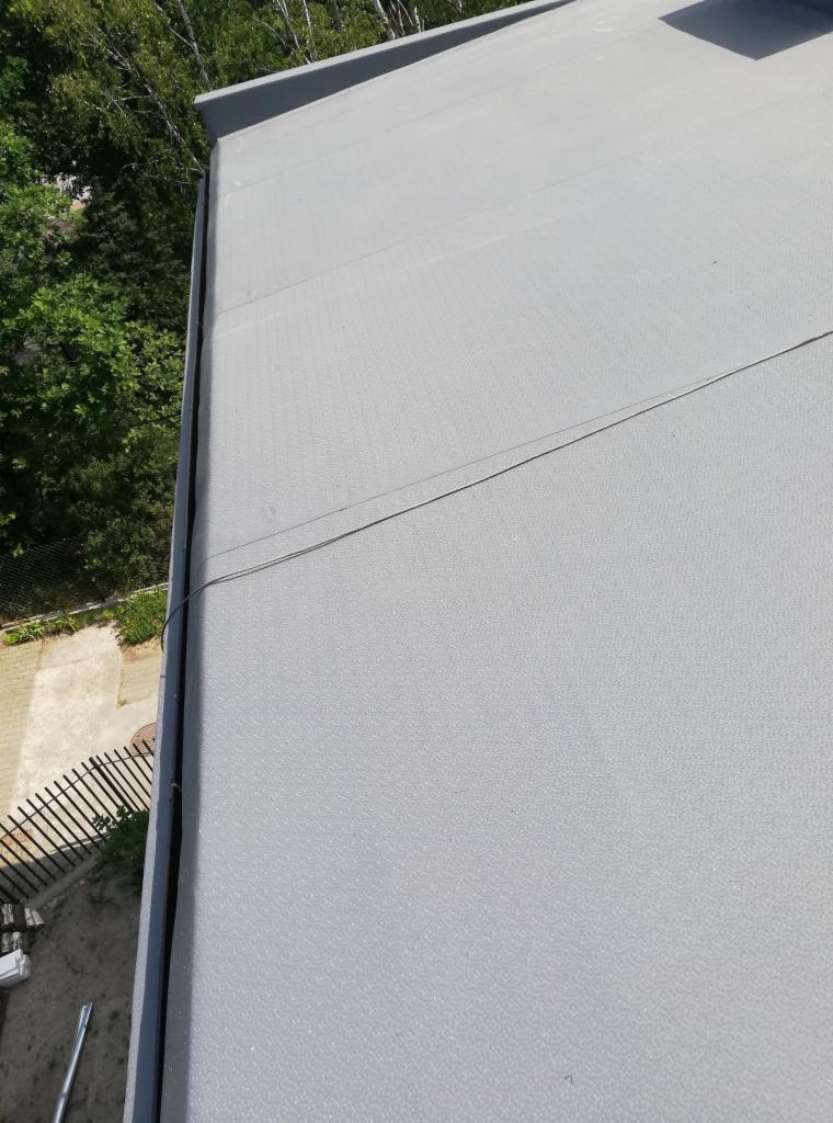 Szary, płaski dach pokryty papą termozgrzewalną z widocznymi łączeniami i fragmentem rynny w tle z zielenią.